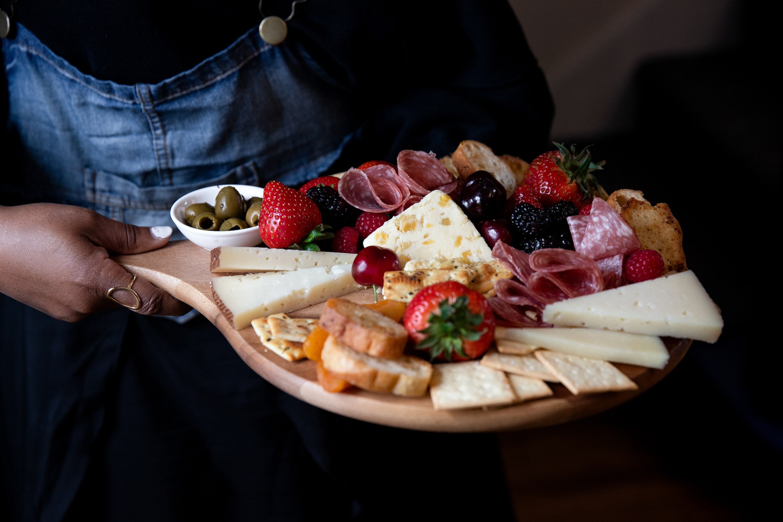 Person holding a charcuterie board with various foods including cheese, fruits, and meats.