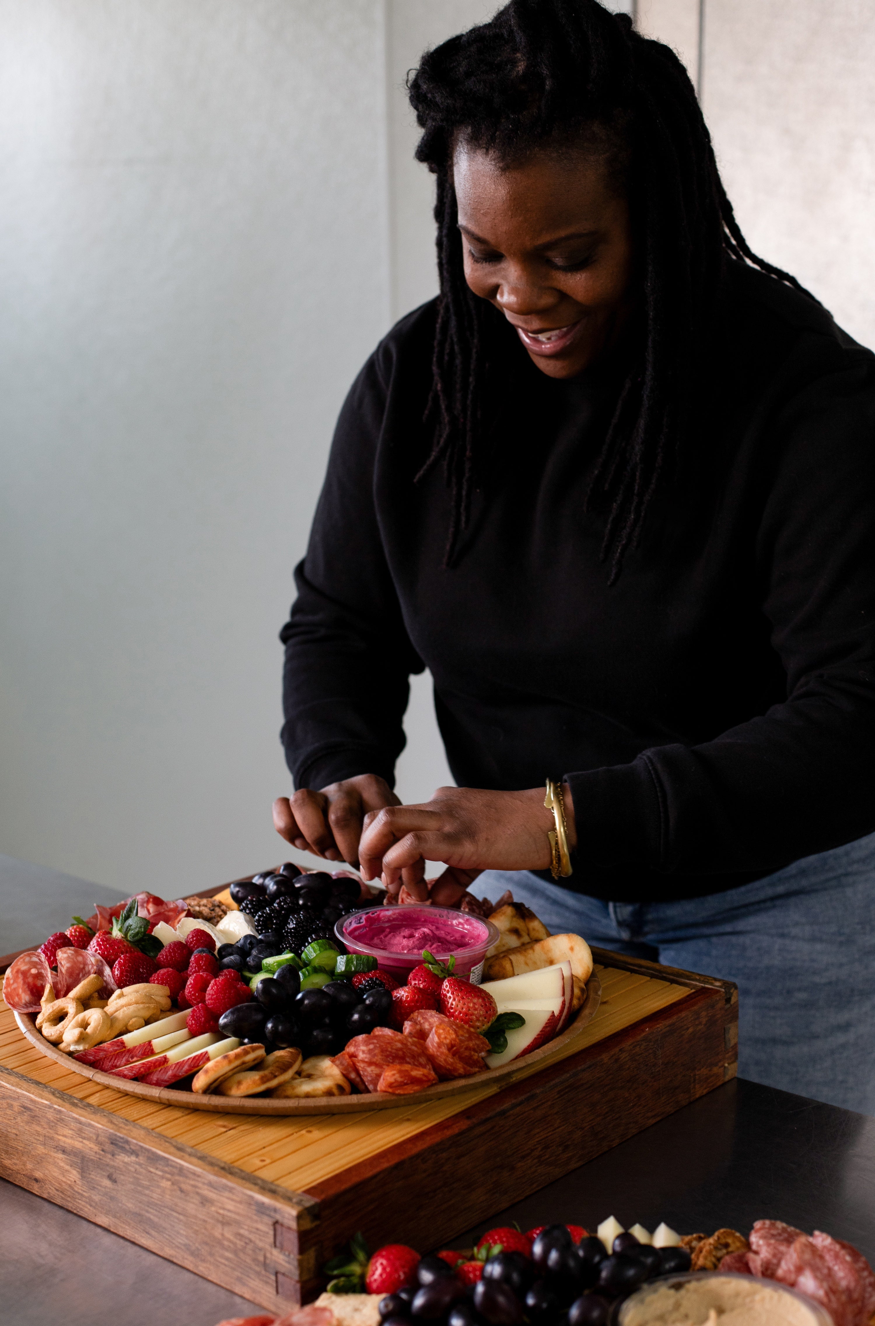 Person preparing a charcuterie board with various fruits and meats on a wooden board.